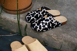 Three clog-style mule slippers arranged outdoors on a dark wooden bench and concrete ledge: one pair is plain light beige suede-like, and the pair above is black and white animal-print with tan soles.