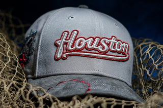 A gray baseball cap with 'Houston' embroidered in red and white script on the front, styled on a bed of fishing net over a blue surface against a dark background.