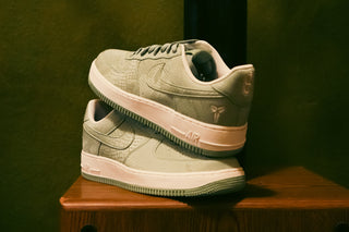 A pair of light sage green and white Nike Air Force 1 Low sneakers stacked on top of a vintage wooden speaker, with a black vase in the background against a dark olive green wall.